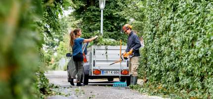 Eine Frau und ein Mann beladen einen Anhänger mit Grünschnitt.