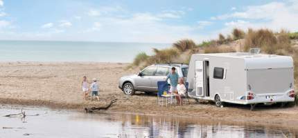 Familie mit Wohnwagen und Auto am Strand, zwei Kinder spielen am Wasser.