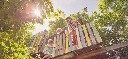 Zwei Kinder und ihr Vater streichen ein Baumhaus in bunten Farben.