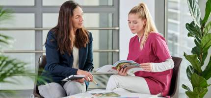 Eine Frau zeigt einer jungen Pflegerin eine Broschüre