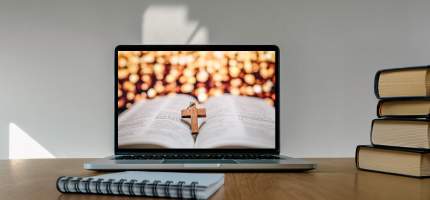 Ein Laptop auf dessen Monitor eine geöffnete Bibel mit einem darauf liegendem kleinen Kreuz aus Holz zu sehen ist.