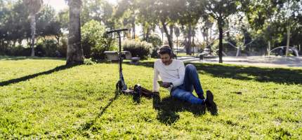 Ein Mann liegt mit Kopfhörern auf einer Wiese im Park neben seinem E-Roller.