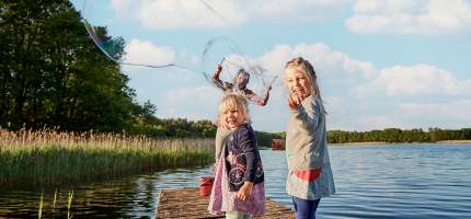 Zwei Kinder und ein Mann stehen auf einem Steg am See und machen Seifenblasen.