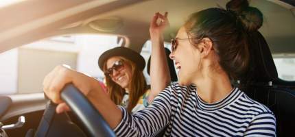 Zwei junge Frauen tragen Sonnenbrillen und singen gemeinsam im Auto.