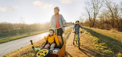 Eine Frau fährt ein Lastenrad mit Ihrem Jungen vorne. Ein anderer fährt mit dem Fahrrad dahinter.