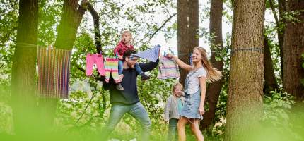 Eine lachende junge Familie hängt Wäsche auf eine Leine zwischen zwei Bäumen.