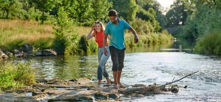 Ein Mann und eine Frau laufen barfuß über Steine in einem Fluss.