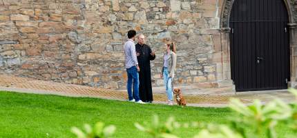 Ein Mann und eine Frau sprechen mit einem Pfarrer vor einer Kirche mit dem Hund an der Leine.