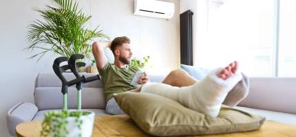 Ein junger Mann mit eingegipsten Fuß sitzt mit seinem Handy auf einer Couch.