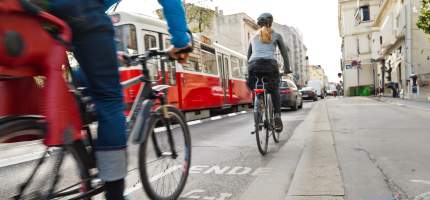 Zwei Fahrradfahrer auf dem Radweg. Es fahren eine S-Bahn und Autos neben ihnen.