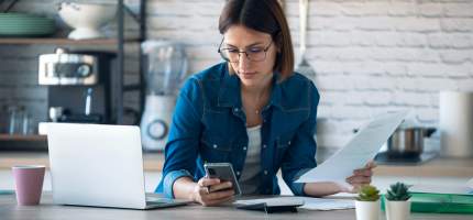 Eine Frau arbeitet an einem Laptop und schaut auf ihr Smartphone.