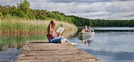 Eine Frau sitzt auf einem Steg und liest während weiterer Personen mit einem Boot auf dem See fahren.