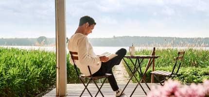 Ein Mann sitzt auf der Terrasse und liest ein Buch. Im Hintergrund liegt ein See.