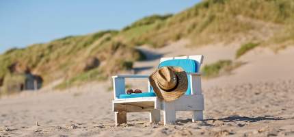 Ein Holzstuhl mit Sitzkissen, Hut und Sonnenbrille steht am Strand.