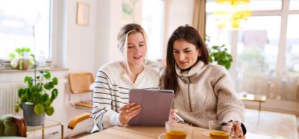 Zwei Frauen trinken Tee und sitzen an einem Tisch und schauen interessiert auf ein Tablet.