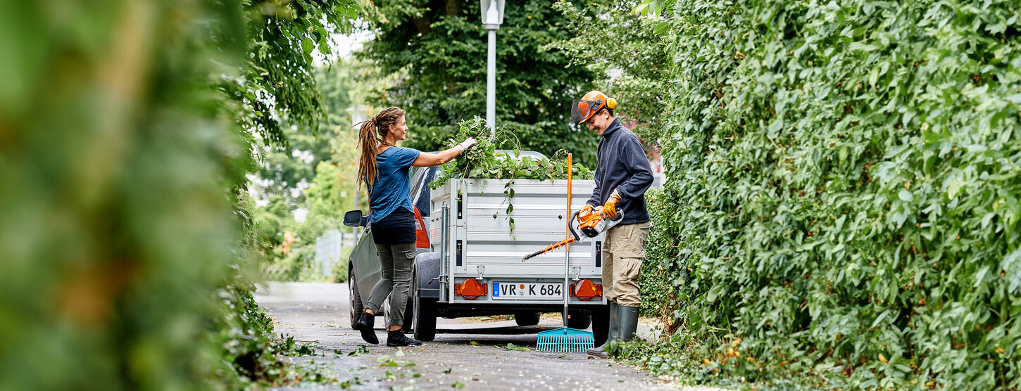 Eine Frau und ein Mann beladen einen Anhänger mit Grünschnitt.