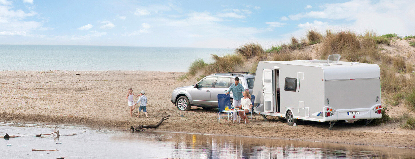 Familie mit Wohnwagen und Auto am Strand, zwei Kinder spielen am Wasser.