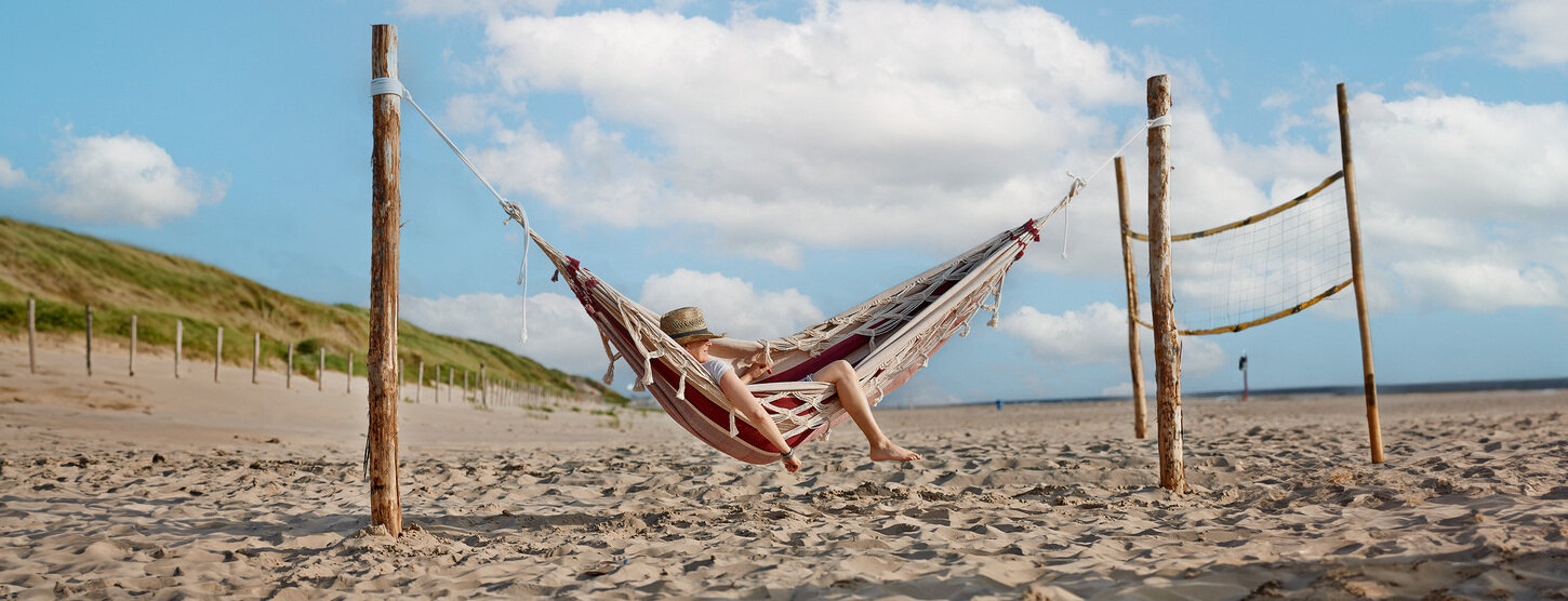 Eine Person mit Hut liegt in einer Hängematte am Strand.