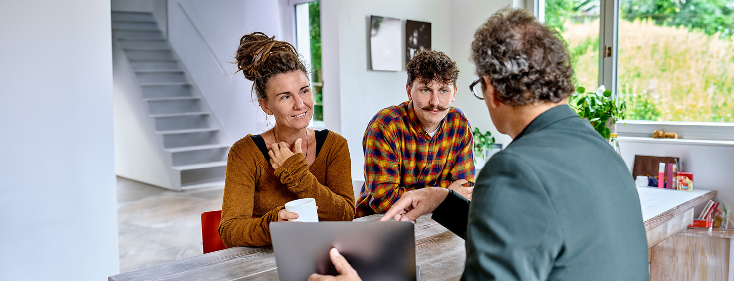 Zwei Personen werden zuhause beraten. Ein Mann zeigt etwas auf seinem Laptop.