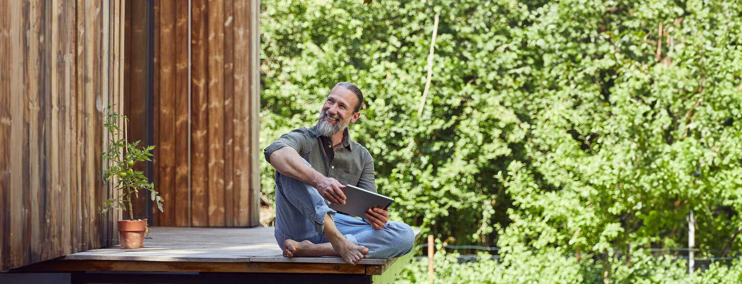 Mann sitzt barfuß auf einer Terrasse aus Holz im Garten.