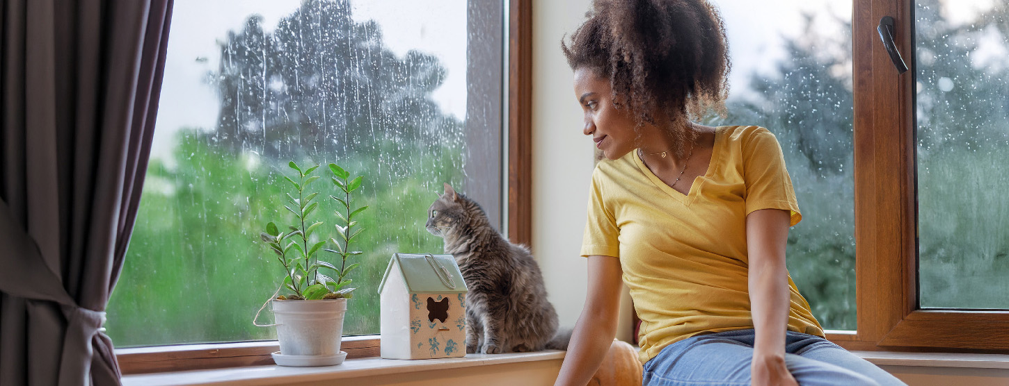 Eine junge Frau schaut mit ihrer Katze bei Regen aus dem Fenster.