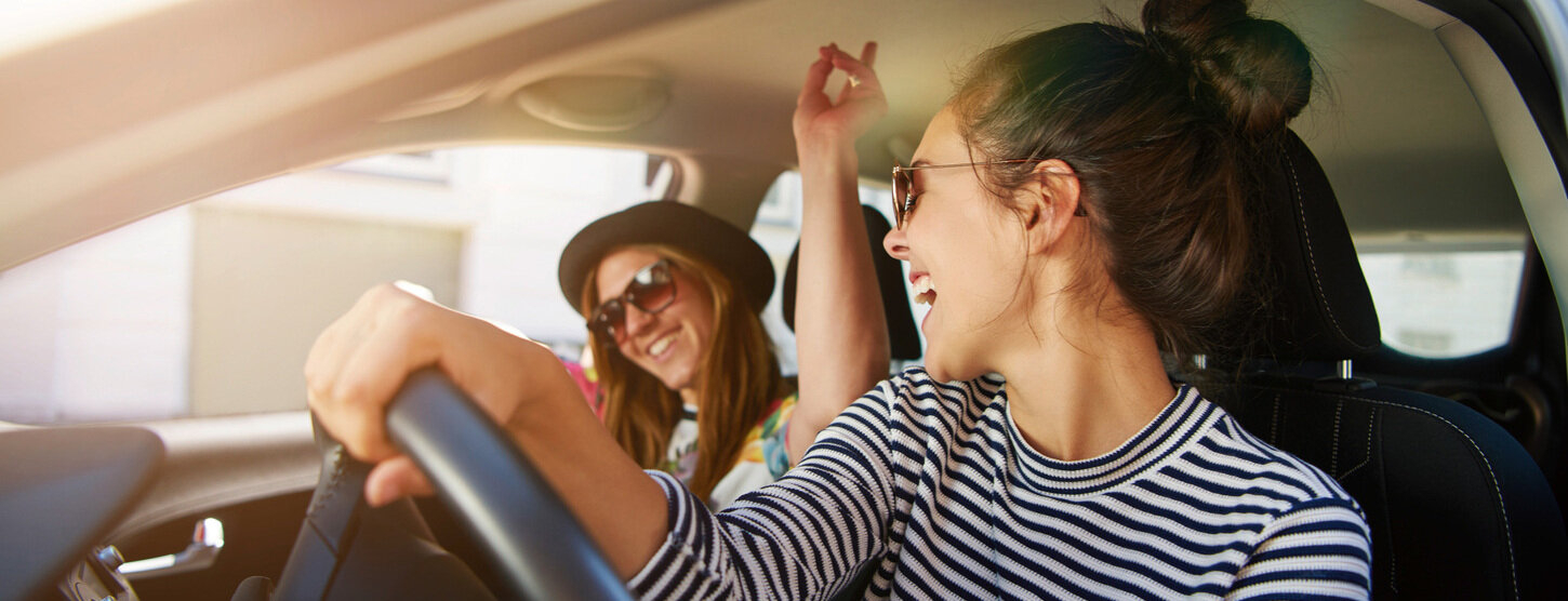 Zwei junge Frauen tragen Sonnenbrillen und singen gemeinsam im Auto.