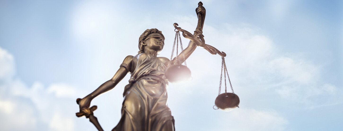 Justitia Bronzefigur vor blauem Himmel mit Wolken.
