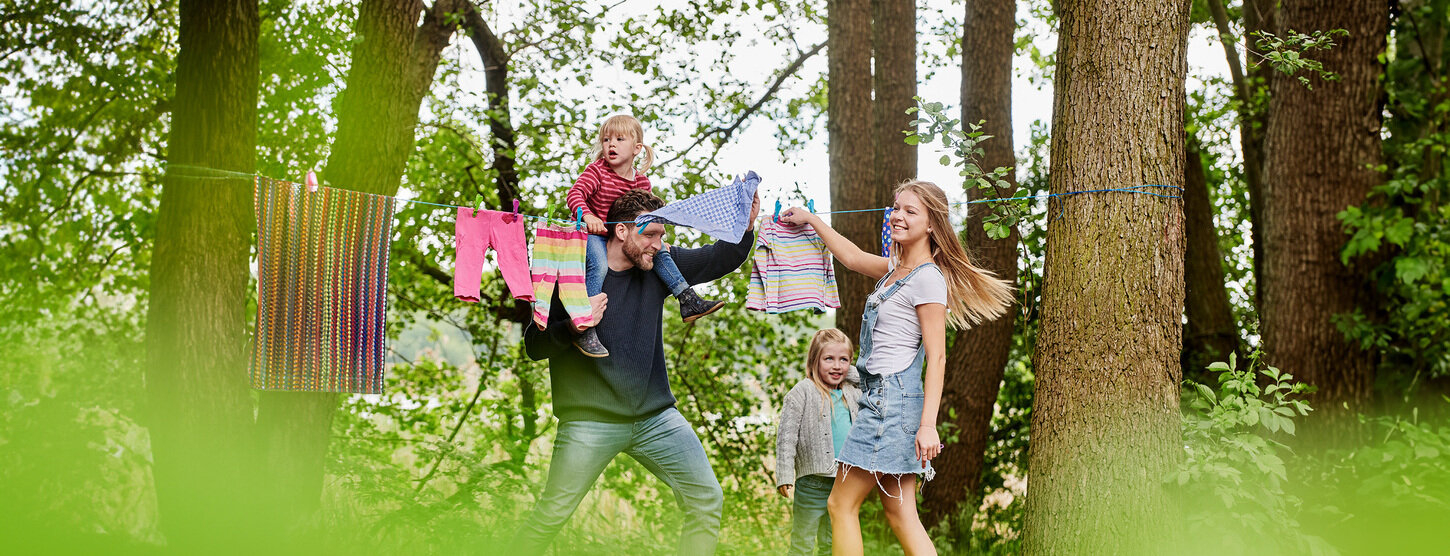 Eine lachende junge Familie hängt Wäsche auf eine Leine zwischen zwei Bäumen.
