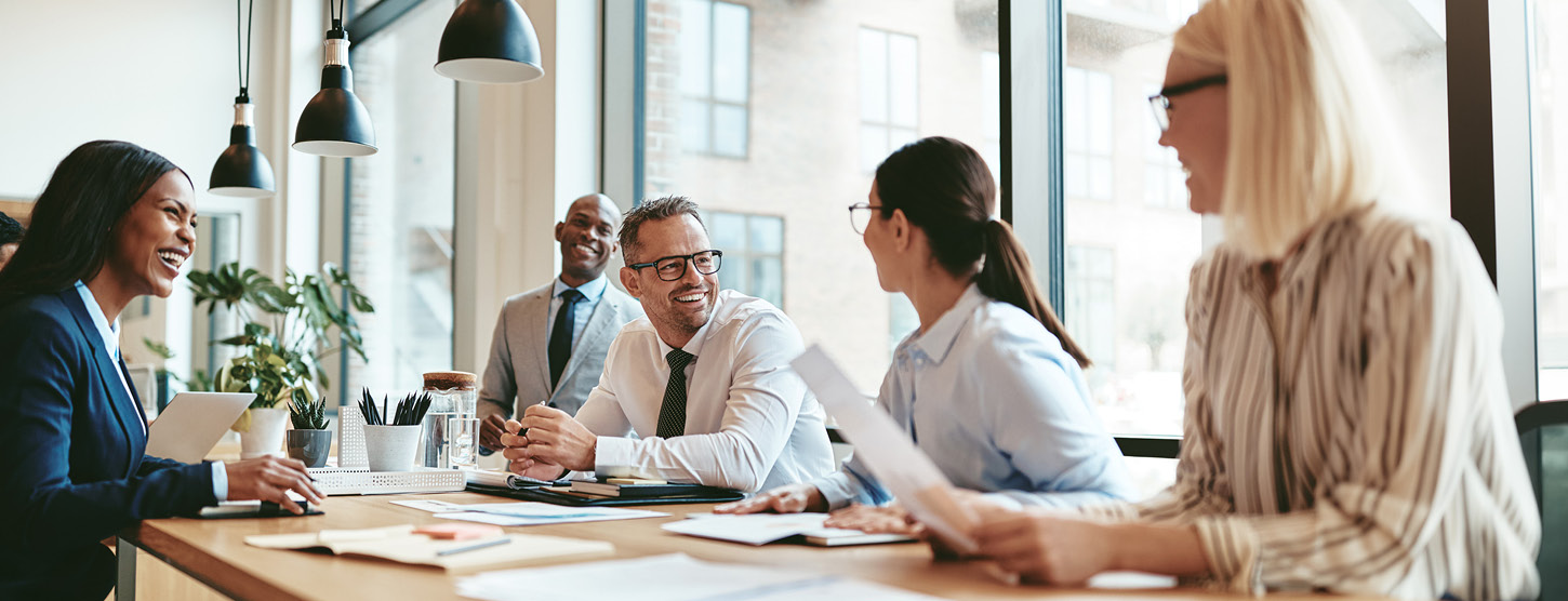 Fünf Personen sitzen lachend an einem Tisch, vor ihnen liegen Stifte und Zettel.