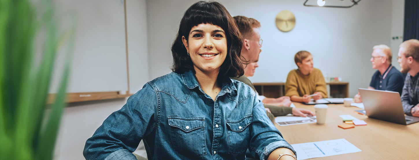 Eine lächelnde Frau sitzt mit anderen Personen an einem Besprechungstisch.