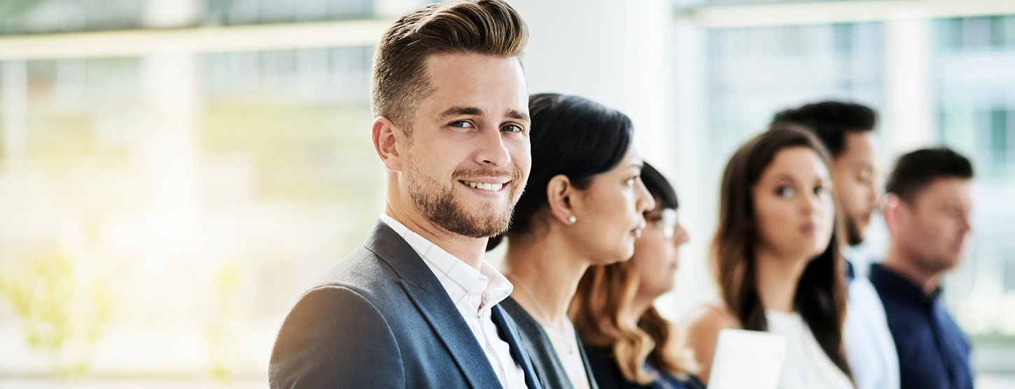 Menschen stehen in einer Reihe. Ein Mann vorne schaut in die Kamera.