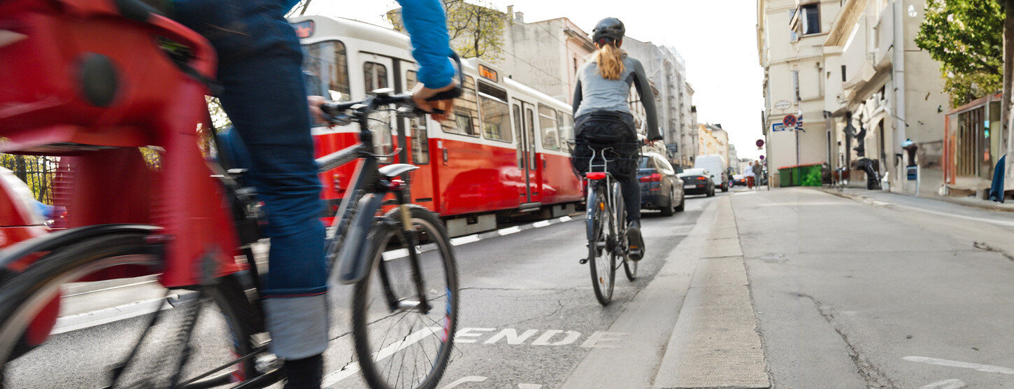 Zwei Fahrradfahrer auf dem Radweg. Es fahren eine S-Bahn und Autos neben ihnen.