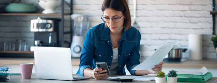 Eine Frau arbeitet an einem Laptop und schaut auf ihr Smartphone.