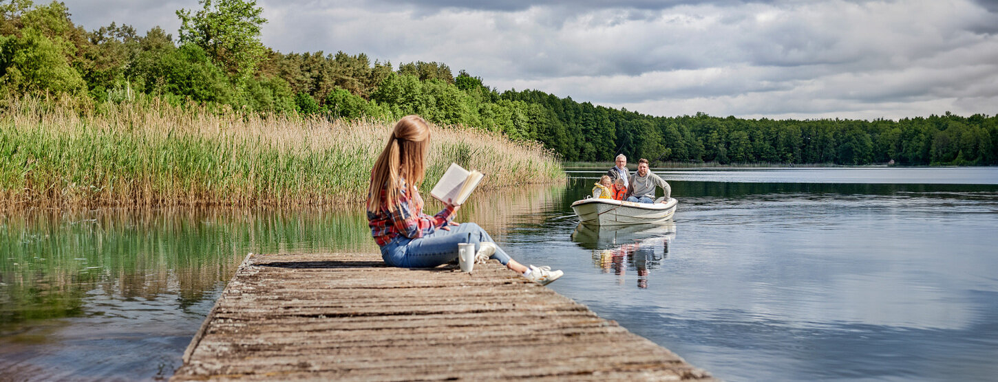 Eine Frau sitzt auf einem Steg und liest während weiterer Personen mit einem Boot auf dem See fahren.