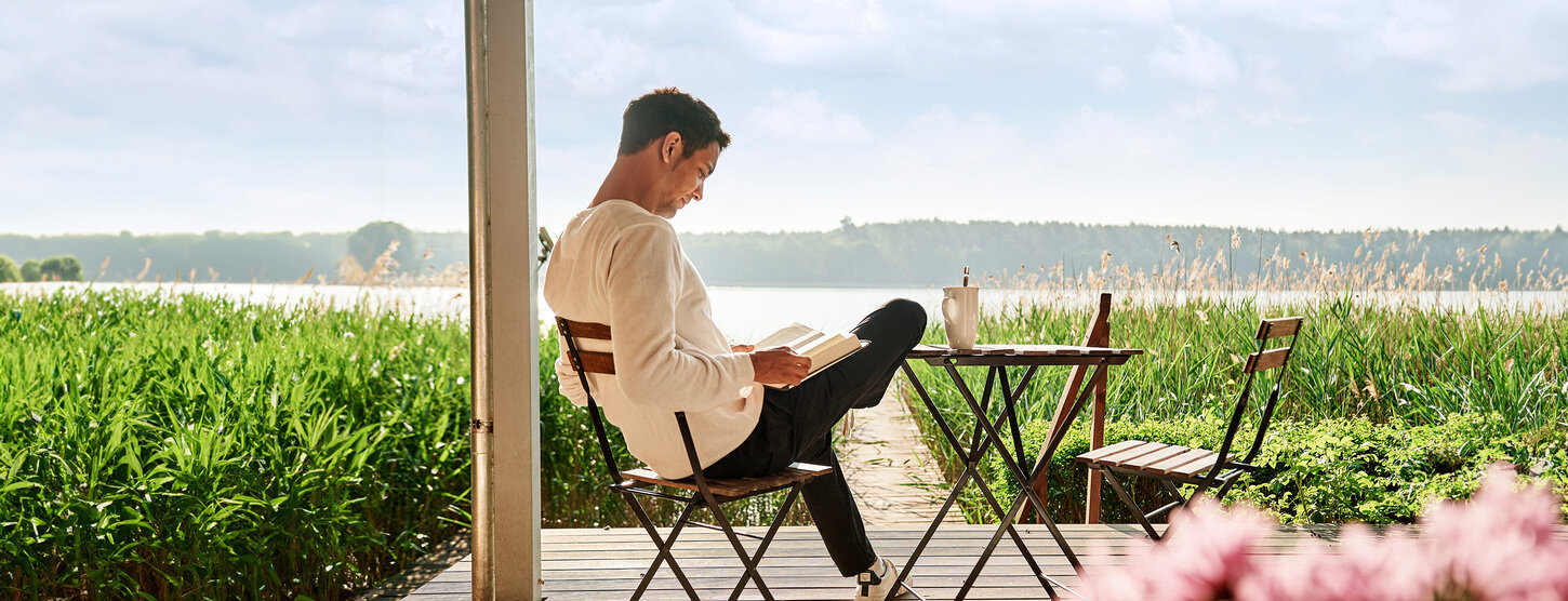 Ein Mann sitzt auf der Terrasse und liest ein Buch. Im Hintergrund liegt ein See.