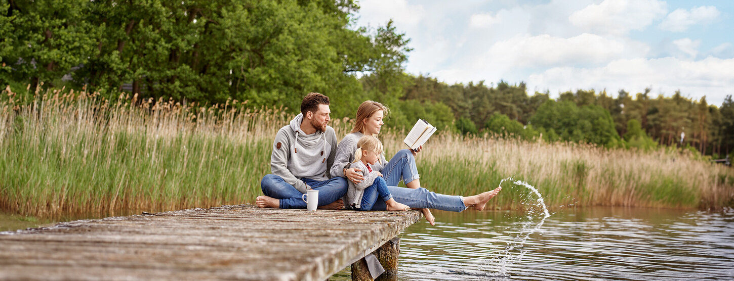 Eine Familie sitzt gemeinsam auf einem Steg und hält die Füße in den See.