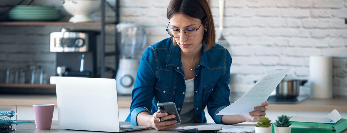 Eine Frau arbeitet an einem Laptop und schaut auf ihr Smartphone.
