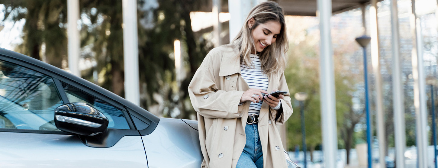 Frau lehnt an Auto und schaut auf ihr Handy.