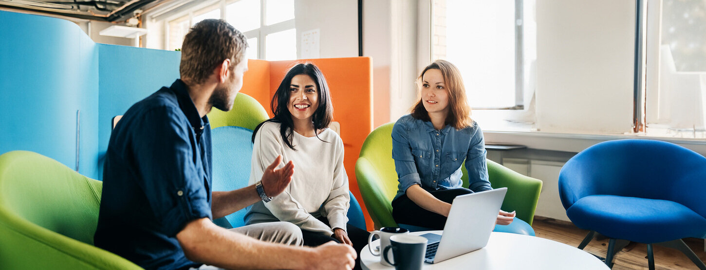 Drei Personen sitzen auf bunten Stühlen, unterhalten sich vor einem Laptop.