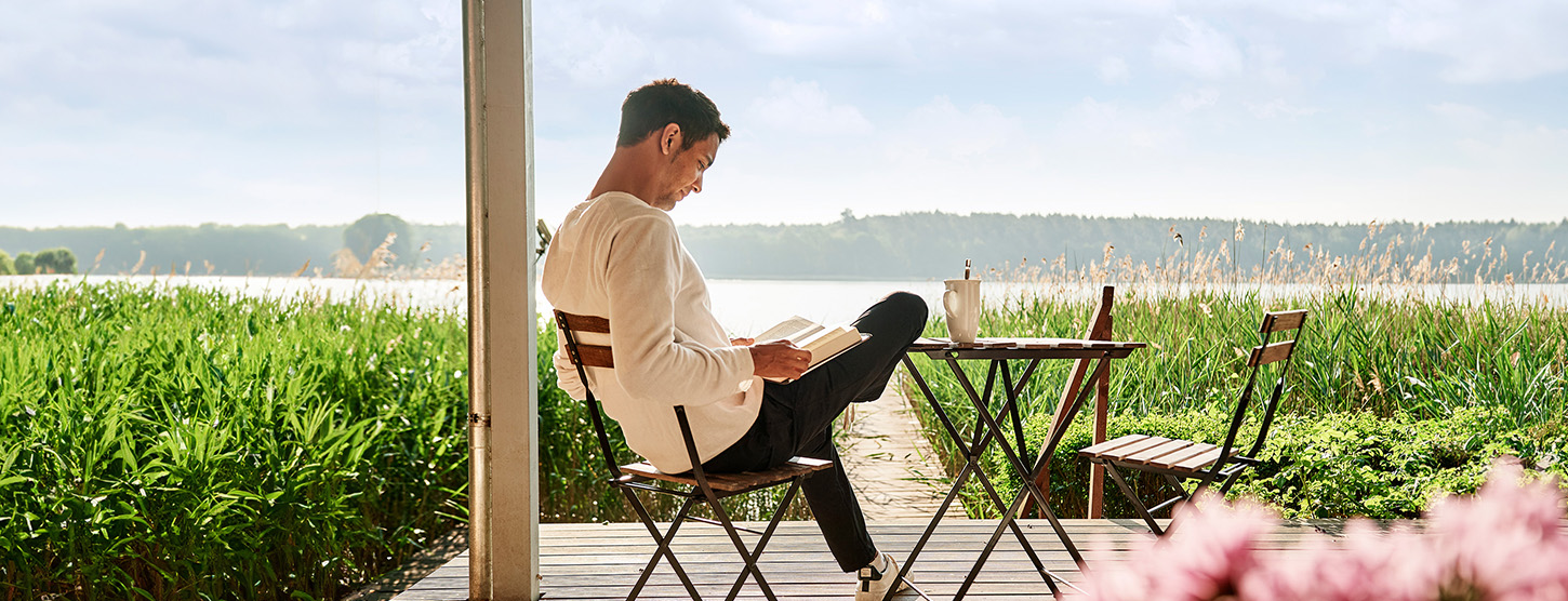 Ein Mann sitzt auf der Terrasse und liest ein Buch. Im Hintergrund liegt ein See.