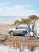 Familie mit Wohnwagen und Auto am Strand, zwei Kinder spielen am Wasser.