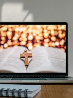 Ein Laptop auf dessen Monitor eine geöffnete Bibel mit einem darauf liegendem kleinen Kreuz aus Holz zu sehen ist.