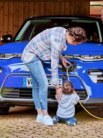 Eine Frau lädt ihr Elektroauto auf. Ihr kleines Kind hilft und hält das Kabel.