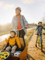 Eine Frau fährt ein Lastenrad mit Ihrem Jungen vorne. Ein anderer fährt mit dem Fahrrad dahinter.