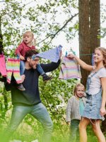 Eine lachende junge Familie hängt Wäsche auf eine Leine zwischen zwei Bäumen.