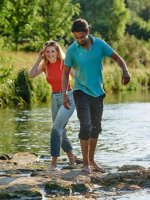 Ein Mann und eine Frau laufen barfuß über Steine in einem Fluss.