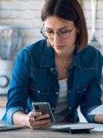 Eine Frau arbeitet an einem Laptop und schaut auf ihr Smartphone.