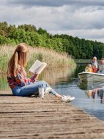 Eine Frau sitzt auf einem Steg und liest während weiterer Personen mit einem Boot auf dem See fahren.