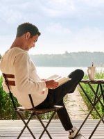 Ein Mann sitzt auf der Terrasse und liest ein Buch. Im Hintergrund liegt ein See.