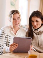 Zwei Frauen trinken Tee und sitzen an einem Tisch und schauen interessiert auf ein Tablet.